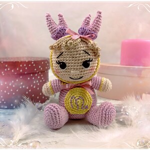 May include: A crocheted amigurumi doll representing the Virgo zodiac sign. The doll is wearing a pink outfit with a yellow star and the symbol for Virgo. The doll has a crown of crocheted flowers on its head.