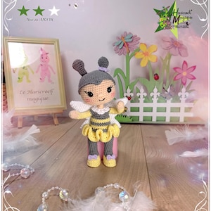May include: A handmade crochet bee doll with a gray head, yellow and black striped body, and yellow skirt. The doll has white wings and purple accents. The doll is standing in front of a decorative garden scene with flowers and a white picket fence.