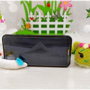 Op de afbeelding: Een groene, gehaakte rups telefoonstandaard met een witte, gehaakte telefoonhouder. De rups heeft roze bloemen op zijn kop en zwarte ogen. De telefoonstandaard staat op een houten oppervlak. De tekst "Les Mysti Croch'" is zichtbaar op de achtergrond.