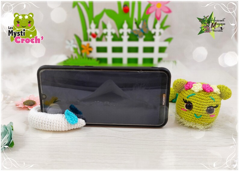 Op de afbeelding: Een groene, gehaakte rups telefoonstandaard met een witte, gehaakte telefoonhouder. De rups heeft roze bloemen op zijn kop en zwarte ogen. De telefoonstandaard staat op een houten oppervlak. De tekst "Les Mysti Croch'" is zichtbaar op de achtergrond.