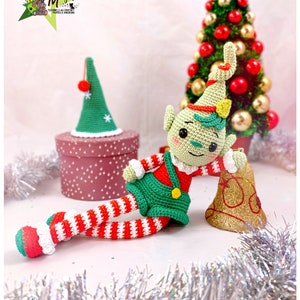 Puede incluir: Un muñeco de elfo de crochet verde con piernas rayadas rojas y blancas está sentado junto a una campana dorada. El elfo lleva un atuendo verde y rojo y un gorro verde con un pompón rojo. El elfo se apoya en la campana con su brazo derecho. El elfo está sentado sobre una superficie blanca con una guirnalda plateada en primer plano. Hay un pequeño árbol de Navidad en el fondo.