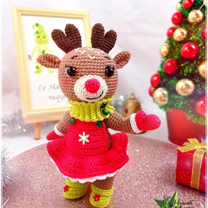 Könnte beinhalten: Häkelpuppe eines Rentiers, die ein rotes Kleid mit einer weißen Schneeflocke, einen grünen Pullover und grüne Stiefel trägt. Die Puppe hat einen braunen Körper, eine rosa Nase und schwarze Augen.