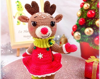 May include: Crocheted reindeer doll wearing a red dress with a white snowflake, green sweater, and green boots. The doll has a brown body, a pink nose, and black eyes.