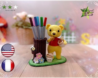Może przedstawiać: Pluszowy miś Winnie the Pooh wykonany na szydełku trzymający słoik miodu, stojący obok brązowego stojaka na ołówki w kształcie pnia drzewa z kolorowymi kwiatami i pszczołą. Miś jest żółty w czerwonej koszulce i z brązowymi oczami. Stojak na ołówki ma zieloną podstawę.