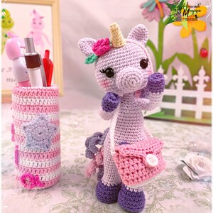 Peut inclure: Une licorne en crochet de couleur violette avec une couronne de fleurs roses et un sac &agrave; main rose. La licorne tient un petit bouton blanc dans sa main. La licorne est assise sur une surface blanche avec un porte-crayons en crochet ray&eacute; rose et blanc.