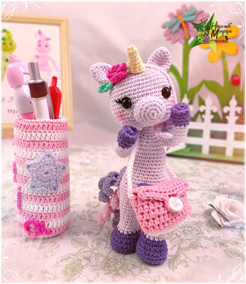 Peut inclure: Une licorne en crochet de couleur violette avec une couronne de fleurs roses et un sac &agrave; main rose. La licorne tient un petit bouton blanc dans sa main. La licorne est assise sur une surface blanche avec un porte-crayons en crochet ray&eacute; rose et blanc.