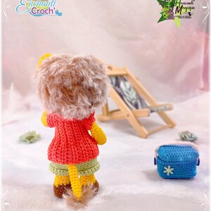 May include: A crocheted lion doll wearing a red jacket and green shorts. The doll is standing on a white surface with a wooden beach chair and a blue crocheted bag in the background.