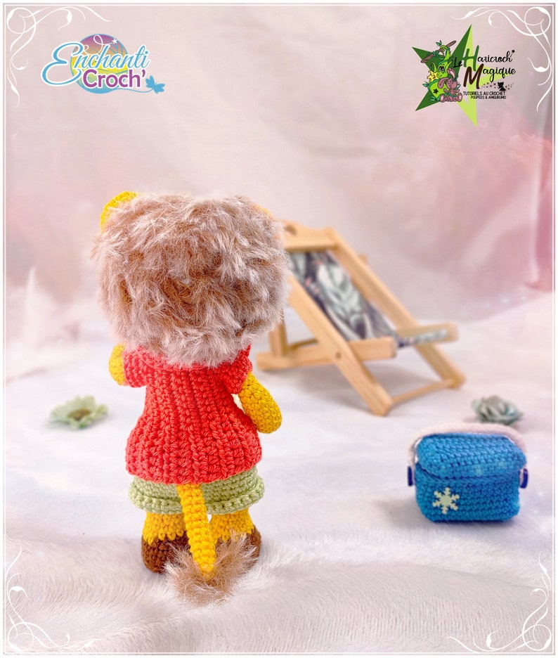 May include: A crocheted lion doll wearing a red jacket and green shorts. The doll is standing on a white surface with a wooden beach chair and a blue crocheted bag in the background.