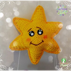 Pode incluir: Uma estrela amarela de croch&ecirc; com um rosto sorridente, olhos pretos e bochechas vermelhas. A estrela est&aacute; pendurada em um barbante.