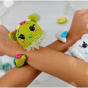 Op de afbeelding: Twee gebreide dierenarmbanden, een groene met roze en blauwe accenten en een witte met blauwe accenten. De armbanden worden gedragen op de polsen van een persoon.