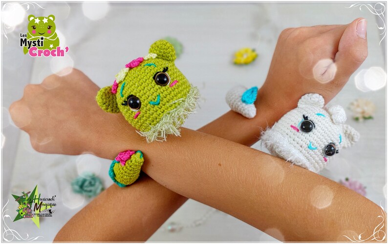 Op de afbeelding: Twee gebreide dierenarmbanden, een groene met roze en blauwe accenten en een witte met blauwe accenten. De armbanden worden gedragen op de polsen van een persoon.