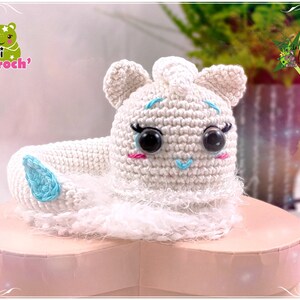 Op de afbeelding: Witte gehaakte unicorn amigurumi speelgoed met blauwe ogen en een blauwe hoorn. De unicorn zit op een roze hartvormige doos.