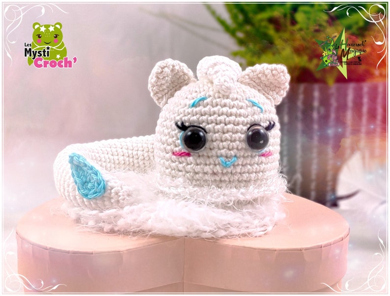 Op de afbeelding: Witte gehaakte unicorn amigurumi speelgoed met blauwe ogen en een blauwe hoorn. De unicorn zit op een roze hartvormige doos.