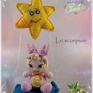 Puede incluir: Una muñeca amigurumi de crochet que representa el signo del zodíaco Escorpio, vestida con un atuendo rosa y morado y sentada en un columpio azul. La muñeca está suspendida de una estrella amarilla con una cara sonriente.