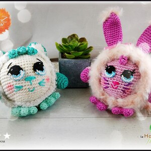May include: Two crocheted stuffed animals, one white with teal accents and the other pink with a fluffy white fur trim. Both have large blue eyes and a pink nose. The white animal has a cat-like face and the pink animal has a bunny-like face.