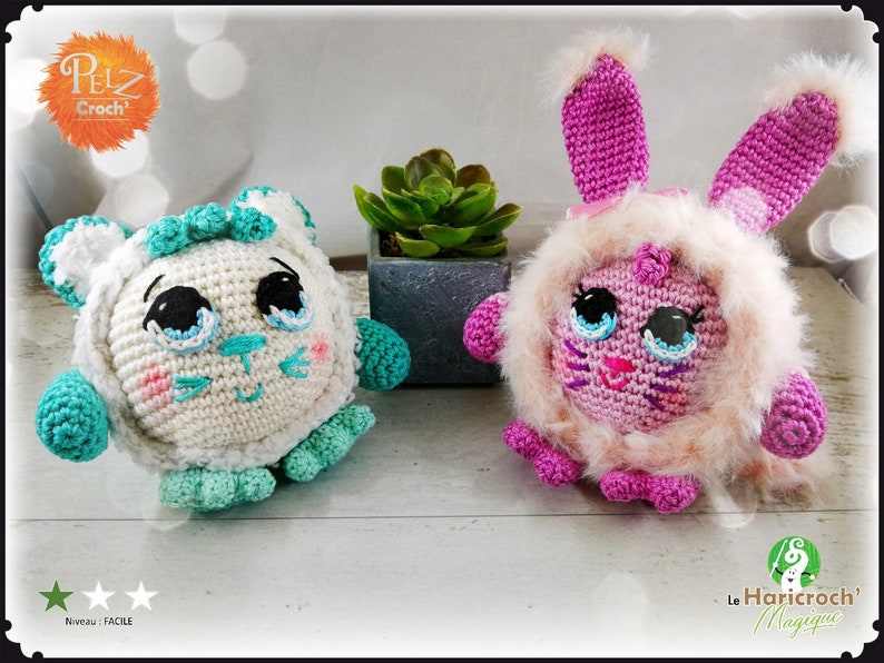 May include: Two crocheted stuffed animals, one white with teal accents and the other pink with a fluffy white fur trim. Both have large blue eyes and a pink nose. The white animal has a cat-like face and the pink animal has a bunny-like face.