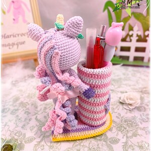 Peut inclure: Un porte-crayons en crochet en forme de licorne avec un design ray&eacute; rose, violet et jaune. La licorne a une crini&egrave;re et une queue rose et violette. Le porte-crayons est rempli de stylos et de crayons.