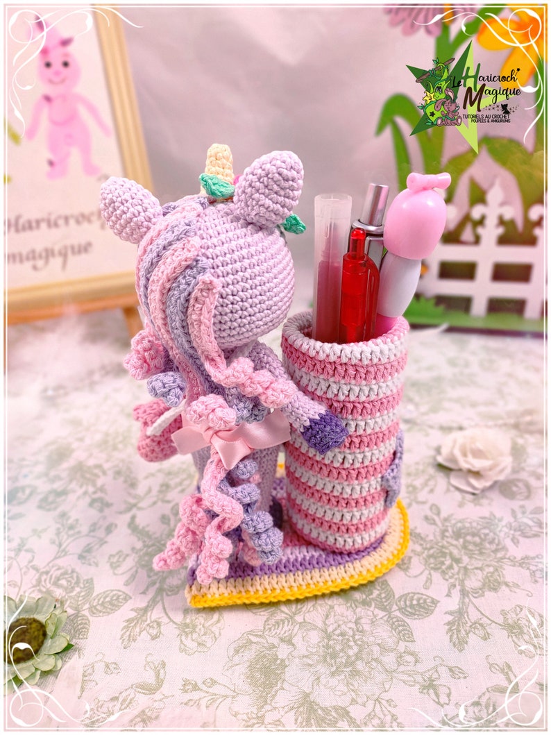 Peut inclure: Un porte-crayons en crochet en forme de licorne avec un design ray&eacute; rose, violet et jaune. La licorne a une crini&egrave;re et une queue rose et violette. Le porte-crayons est rempli de stylos et de crayons.