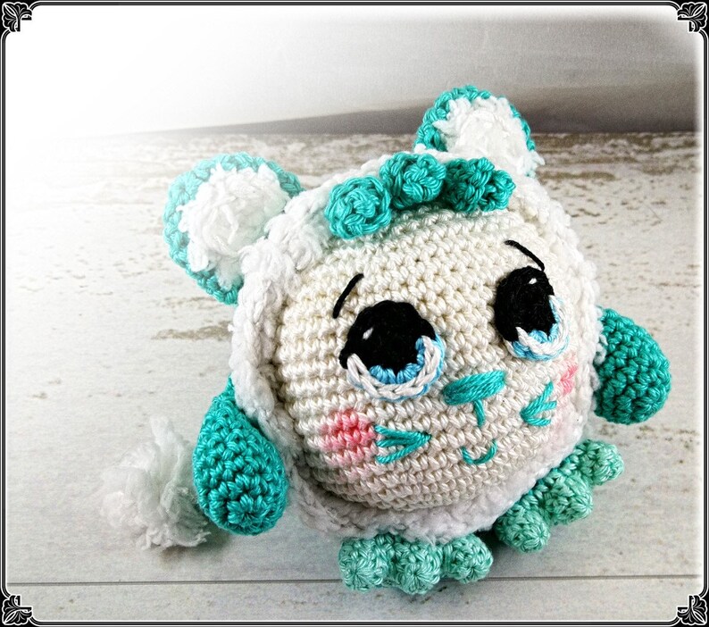 May include: A crocheted stuffed animal with a white body, teal blue accents, and a cute face. The animal has large eyes, a small nose, and a stitched smile.