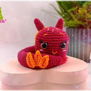 Op de afbeelding: Een gehaakt amigurumi-speelgoed in de vorm van een rode draak met oranje klauwen. De draak zit op een roze hartvormige doos. Het speelgoed is handgemaakt en heeft een schattige uitdrukking.