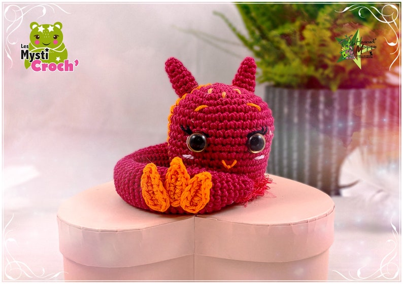Op de afbeelding: Een gehaakt amigurumi-speelgoed in de vorm van een rode draak met oranje klauwen. De draak zit op een roze hartvormige doos. Het speelgoed is handgemaakt en heeft een schattige uitdrukking.