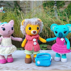 May include: Three crocheted stuffed animals: a pink cat, a yellow lion, and a blue koala. Each animal is wearing a dress and has a sweet expression. The lion is holding a blue crocheted basket.
