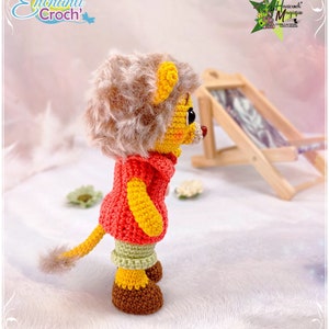 May include: A crocheted lion toy wearing a red and yellow sweater, green pants, and brown shoes. The toy is standing on a white fluffy surface.