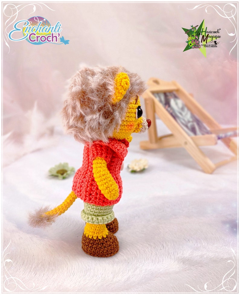 May include: A crocheted lion toy wearing a red and yellow sweater, green pants, and brown shoes. The toy is standing on a white fluffy surface.