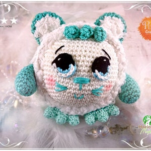 May include: A white crocheted amigurumi toy with teal accents. The toy has large eyes, a small pink nose, and a stitched smile. The toy is sitting on a white fluffy surface.