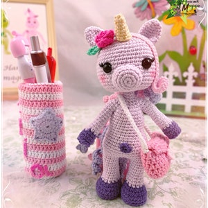 Peut inclure: Un jouet en crochet en forme de licorne de couleur violette avec une fleur rose sur la t&ecirc;te et un sac rose en bandouli&egrave;re. La licorne est debout sur une surface blanche avec un porte-crayons en crochet ray&eacute; rose et blanc en arri&egrave;re-plan.