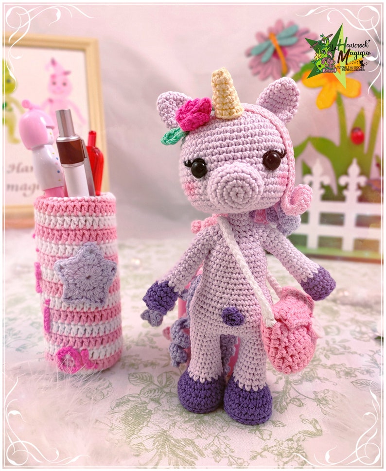 Peut inclure: Un jouet en crochet en forme de licorne de couleur violette avec une fleur rose sur la t&ecirc;te et un sac rose en bandouli&egrave;re. La licorne est debout sur une surface blanche avec un porte-crayons en crochet ray&eacute; rose et blanc en arri&egrave;re-plan.