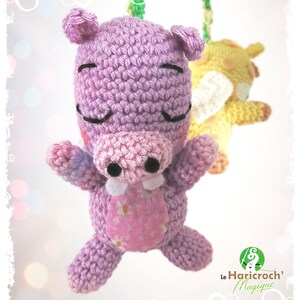 K&ouml;nnte beinhalten: Ein handgefertigtes, geh&auml;keltes lila Hippo-Spielzeug mit rosa Schnauze und Bauch und geschlossenen Augen. Ein gelbes geh&auml;keltes Tier ist im Hintergrund leicht unscharf. Das Nilpferd hat einen rosa Bauch mit Blumenmuster. Das Logo "Le Haricroch' Magique" befindet sich unten rechts.
