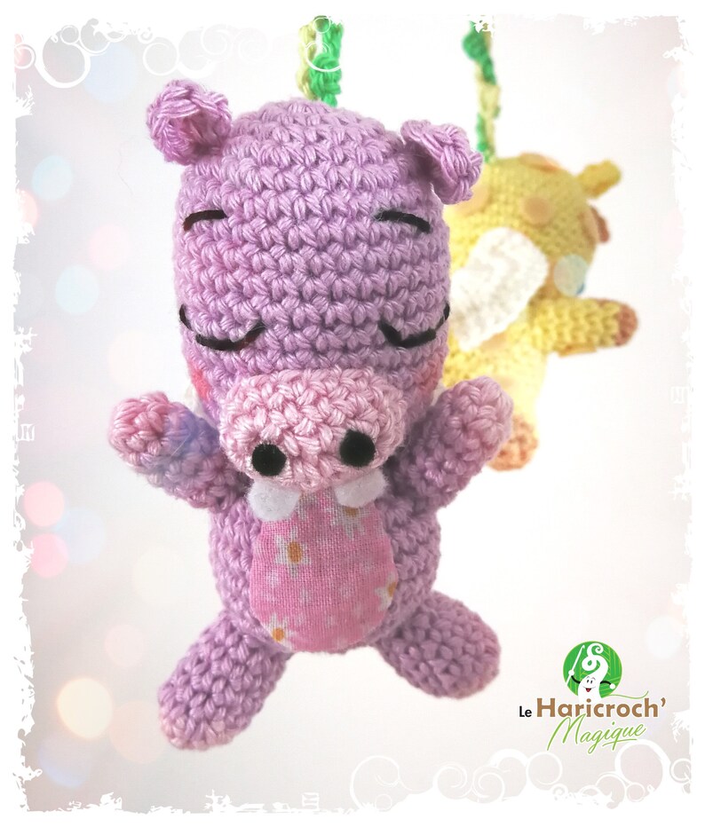 K&ouml;nnte beinhalten: Ein handgefertigtes, geh&auml;keltes lila Hippo-Spielzeug mit rosa Schnauze und Bauch und geschlossenen Augen. Ein gelbes geh&auml;keltes Tier ist im Hintergrund leicht unscharf. Das Nilpferd hat einen rosa Bauch mit Blumenmuster. Das Logo "Le Haricroch' Magique" befindet sich unten rechts.