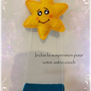 Pode incluir: Uma estrela amarela de croch&ecirc; com um rosto sorridente pendura em um barbante branco com um assento azul de croch&ecirc;. O texto "Inclus la suspension pour votre astro croch" est&aacute; escrito abaixo da estrela.
