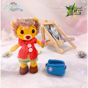 May include: A crocheted lion wearing a red jacket, green shorts, and brown shoes. The lion is holding a small ice cream cone. The lion is sitting on a white surface with a blue crocheted cooler and a wooden beach chair in the background.