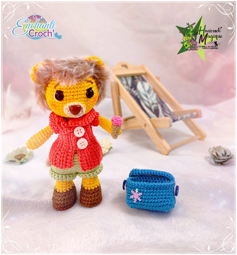 May include: A crocheted lion wearing a red jacket, green shorts, and brown shoes. The lion is holding a small ice cream cone. The lion is sitting on a white surface with a blue crocheted cooler and a wooden beach chair in the background.