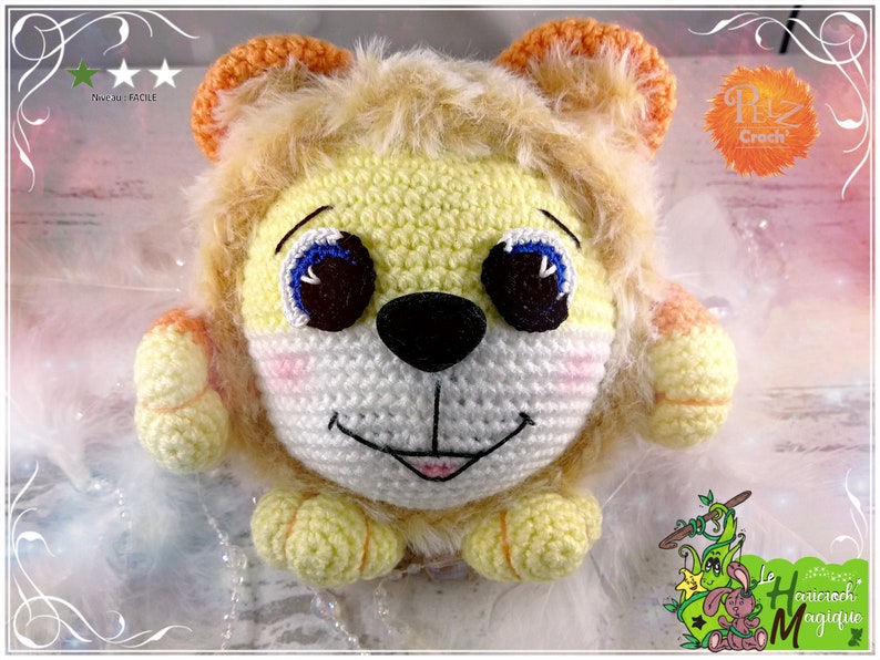 May include: A crocheted lion plush toy with a yellow and orange mane, white face, and black nose. The toy has blue eyes and a pink mouth.
