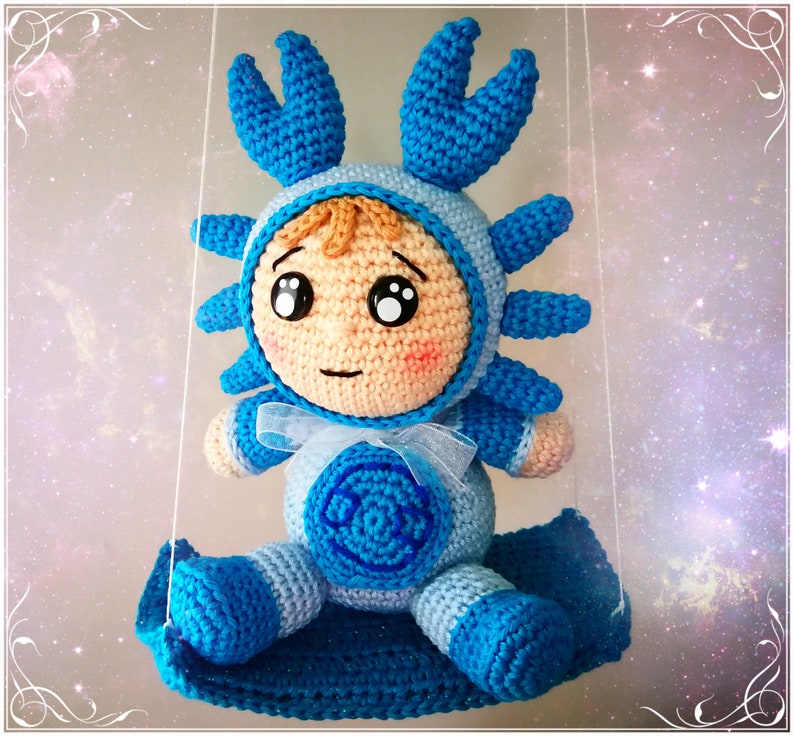 Pode incluir: Um brinquedo de pel&uacute;cia azul e branco de croch&ecirc; em forma de caranguejo. O brinquedo tem um rosto humano com olhos grandes e uma boca pequena. O brinquedo est&aacute; sentado em um balan&ccedil;o de croch&ecirc; azul.