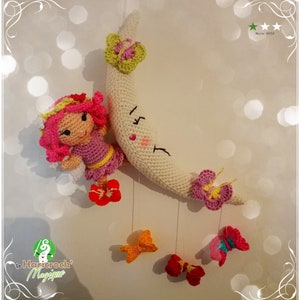Peut inclure: Un mobile en crochet en forme de lune avec une poupée de fée rose, une lune en croissant blanc et cinq papillons colorés suspendus. La lune a un cœur rose et un visage souriant. Le mobile est étiqueté "Le Haricroch' Magique".