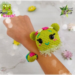 Op de afbeelding: Een groen gehaakt amigurumi-speelgoed met roze en gele bloemen op zijn hoofd. Het speelgoed draagt ​​een groene gehaakte armband met een roze bloem. Het speelgoed zit op de pols van een persoon.