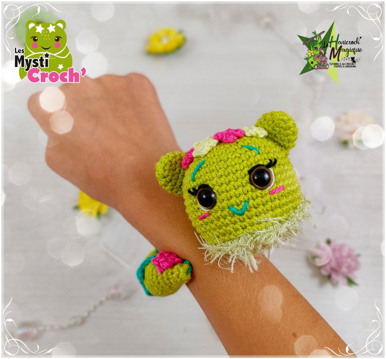 Op de afbeelding: Een groen gehaakt amigurumi-speelgoed met roze en gele bloemen op zijn hoofd. Het speelgoed draagt ​​een groene gehaakte armband met een roze bloem. Het speelgoed zit op de pols van een persoon.