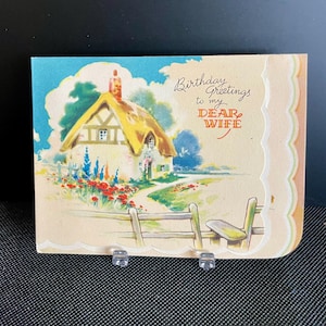 Peut inclure: Carte d'anniversaire vintage avec une illustration à l'aquarelle d'un cottage au toit de chaume, d'une clôture et d'un jardin. La carte porte l'inscription "Birthday Greetings to my DEAR WIFE" en écriture élégante.