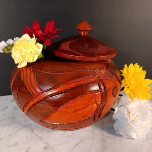 May include: A wooden carved box with a lid, featuring a geometric design in shades of brown and red. The box is decorated with yellow and white flowers.