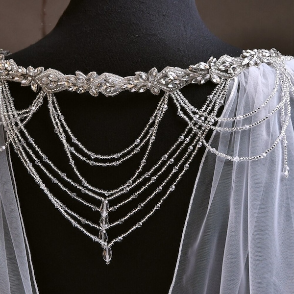 May include: A delicate bridal cape with a beaded and crystal embellishment. The cape is made of sheer tulle and features a beaded and crystal design that drapes down the back.