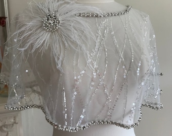 Puede incluir: Una capa blanca de lentejuelas y cuentas con un broche de plumas blancas y diamantes de imitación. La capa tiene un borde festoneado y está adornada con cuentas.