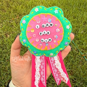 May include: A felt award ribbon featuring a bright green scalloped border and a pink circular center. The text I CAN FIX HIM is spelled out in white plastic letter beads. The design includes small floral sequins and two pink felt tails with white lace.
