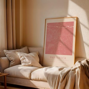 Può includere: Una stampa incorniciata di Marrakech, Marocco, in una calda tonalità rosa, è appesa sopra un divano beige con cuscini e una coperta. La stampa presenta un design a forma di mappa con il nome e le coordinate della città in basso.