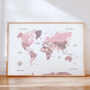 Peut inclure: Une impression encadrée d'une carte du monde dans des tons roses et gris, avec les noms des pays et les étiquettes des océans. La carte comprend une rose des vents et est posée sur un fond blanc. Le cadre est de couleur bois clair.