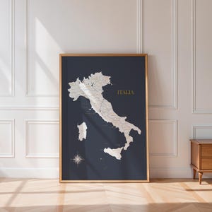 May include: A framed map of Italy, featuring a dark blue backdrop and gold detailing. The map highlights the country's regions in white, with the word "ITALIA" in gold. A compass rose is at the bottom of the map.