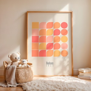 Op de afbeelding: Een ingelijste print met een geometrisch ontwerp in perzik, koraal en geel. De print bevat de tekst "Bauhaus 1925". Een gevlochten mand met twee teddyberen staat voor de print, op een zacht tapijt.